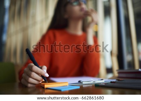 Selective focus on tender manicure and stylish pen which thoughtful female student in red sweatshirt holding in hand.Colored stickers on table.Cropped blurred image of young woman in eyeglasses