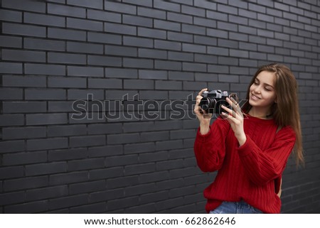 Charming young female tourist dressed in red sweater making photo on vintage camera having city tour on spring day standing on promotional background, hipster girl fond of photography enjoying hobby