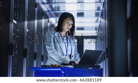 Asian Female IT Engineer Working on a Laptop on Tool Cart, She Scans Hard Drives.  She's in a Big Data Center Full of Rack Servers. Royalty-Free Stock Photo #662881189