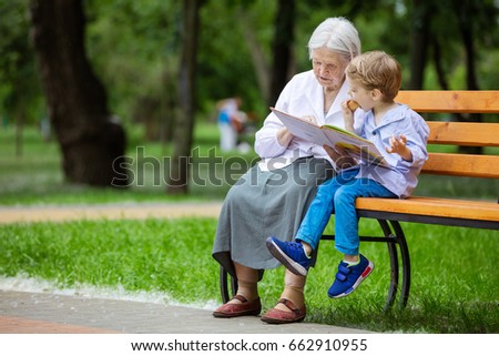 Young boy and great grandmother reading book in summer park, grandson eating bun Royalty-Free Stock Photo #662910955