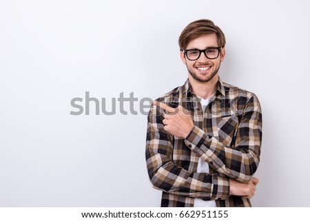 Smiling young nerdy bearded stylish student is standing on pure background in glasses and casual  outfit, pointing on the copyspace