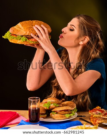 Woman eating french fries and hamburger with pizza. Student consume fast food on table. Girl trying to eat junk on dark background. Cook teaches cook and shares recipes. Girl suffering from gluttony.