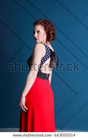 Pretty young woman with hairdo and makeup in long dress looks away in blue studio