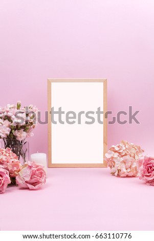 Empty white frame, pink carnations in glass vases, pink roses, beige hydrangea and scented candle on pink background. Sweet and romantic interior. Wedding's background.