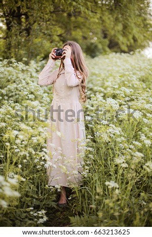 Dreamy relaxed and happy teenager girl taking photo with old camera, walk in the blooming meadow surrounded by white flowers of ground elder in the magic light of sunset in vintage linen dress