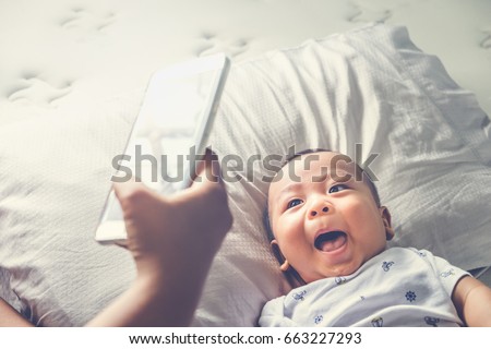 Above high angle shot of happy mom taking photo her Asian baby boy in bed with a smartphone at home. It is a memorable feeling for every parent when their child is growing up. Top down. Overhead view.