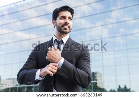 Arabic serious smiling happy successful positive businessman or worker in black suit with beard standing in front of an office glass building and straightens his white shirt with his hand. Royalty-Free Stock Photo #663263149