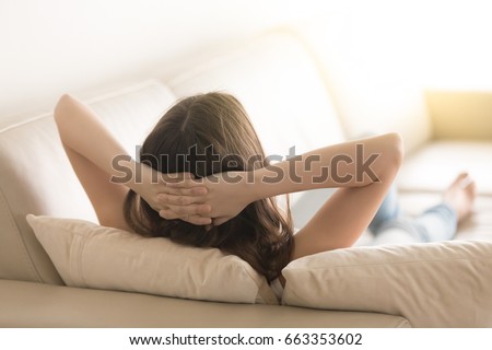 Relaxed woman lying on comfortable sofa on pillows with hands behind head in living room. Carefree lady resting at home or hotel, dreaming about happy future. Day off work concept. Close up. Rear view Royalty-Free Stock Photo #663353602