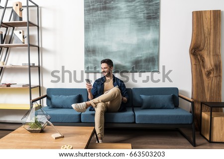 Happy youthful guy bearded resting with cellphone in living room Royalty-Free Stock Photo #663503203