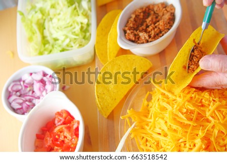 A man puts ground beef into a taco shell with grated cheese, shredded lettuce, chopped tomatoes and onions nearby