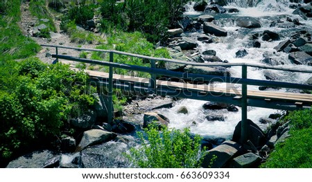 Beautiful view on the high green mountains peaks and a wooden old bridge over the river, blue sky background. Mountain hiking paradise landscape, A stream flows down the rocks, no people.