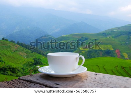 a selective focus picture of a cup of coffee on wooden table beside terraced rice field