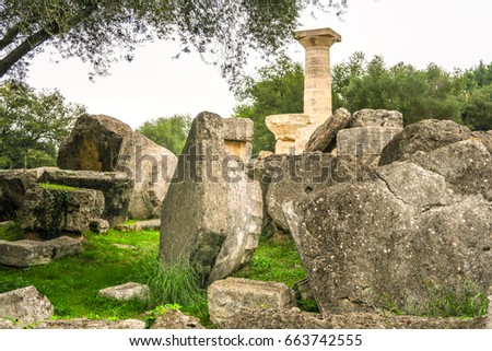 Solitary column standing in background with other column pieces and doric capital laying in foreground on bright green grass and gray sky background