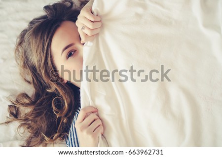 Beautiful young woman lying down in the bed after sleeping. Teen girl with open eyes covers her face with white blanket. Morning concept. View from above with place for your text. Copy space. Royalty-Free Stock Photo #663962731