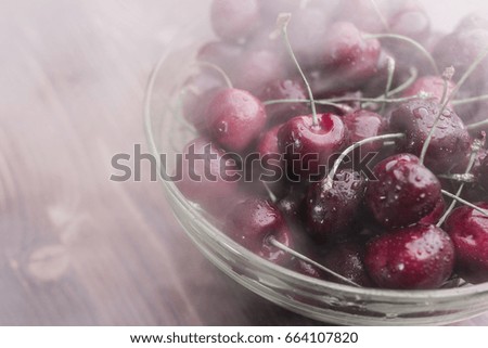 A bright ripe cherry covered with droplets of water lies in a transparent bowl in a cold steam on a brown wooden background