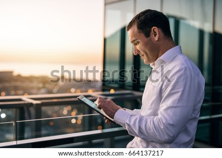 Smiling mature businessman working online with a digital tablet while standing outside on an office building balcony overlooking the city at dusk Royalty-Free Stock Photo #664137217