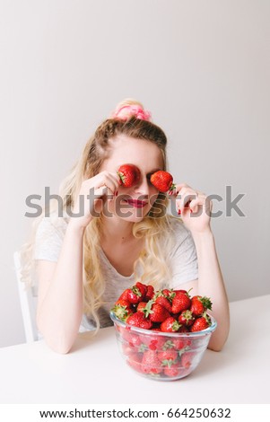 Studio portrait of attractive smiling blond girl sitting on a white wooden chair near a bowl of tasty berries in front of her and putting juicy strawberries on her eyes.
