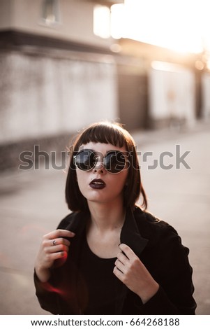 A beautiful young woman with round glasses is posing on the street at sunset