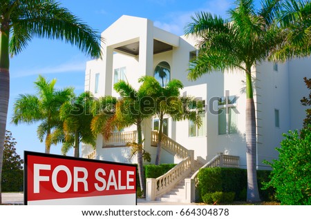 Beautiful luxury residential home with front stairways and lush green palm trees against a blue morning sky with a bright FOR SALE sign in the front yard.