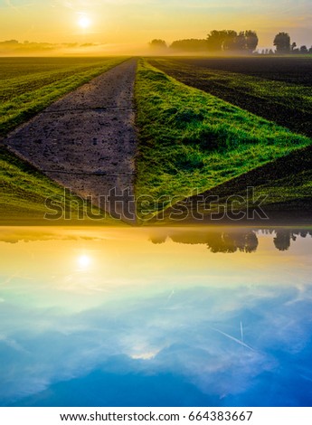Fantasy fine art color image of a foggy autumnal sunrise in a rural landscape with golden light, blue sky, field, trees and a path towards the horizon in a surreal mirrored world
