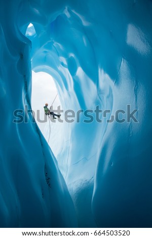 An ice climber in a green jacket and orange helmet rappels past a large rounded entrance to an ice cave on the Matanuska Glacier in Alaska.