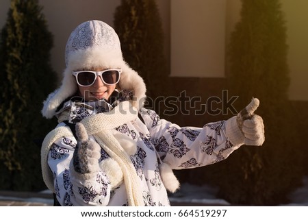 Funny little girl in sunglasses in winter   a sunny day. Thumb up sign. Caucasian stylish little girl in winter clothes walking outdoor. Pretty charming little girl. Portrait of little fashion  girl .