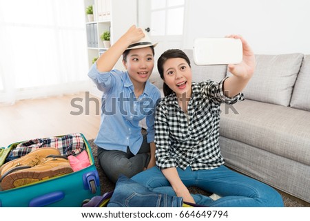 pretty smiling female friends packing luggage at home and using mobile smart phone taking photo selfie trying hat in living room.