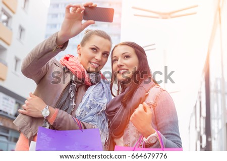 Female friends with shopping bags taking photos through mobile phone