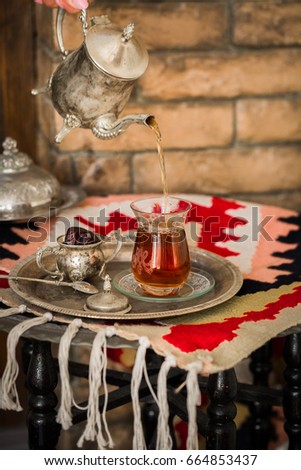 Tea set in oriental style in pear shaped glass with spoon and vintage kettle in female hands poring tea into the glass with dates fruit by side on silver tray on ethnic style rug with tassels.