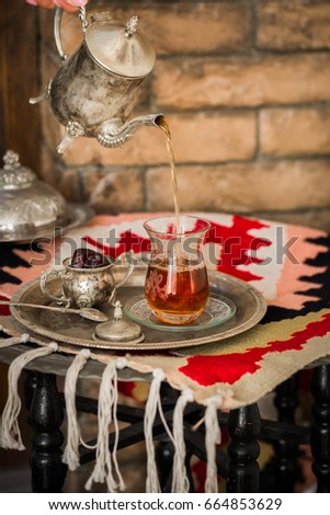Tea set in oriental style in pear shaped glass with spoon and vintage kettle in female hands poring tea into the glass with dates fruit by side on silver tray on ethnic style rug with tassels.