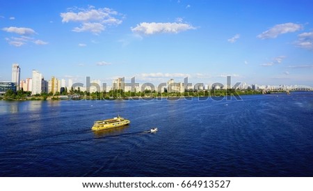 Yellow ship with people goes along Dnieper (Dnepr, Dnipro), Kiev, Ukraine. Beach shore is covered with green bushed and sunbathing people on background of cityscapes, blue  sky with white clouds