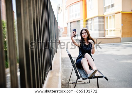 Brunette girl at black dress, sunglasses sitting on bench, listening music from headphones phone, and posing at street of city.