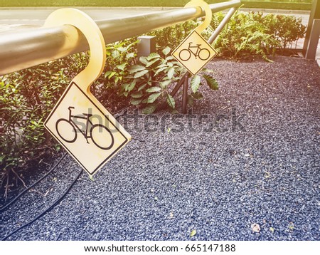Yellow bicycle stop signs are hanged the stainless rail, which is above the gravel ground and plant around.