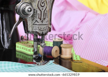  On the old sewing machine lie wooden retro coils with threads, a thimble, a measuring tape and a piece of cotton fabric. Close-up. Retro stylized photo