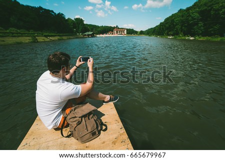 Man sitting on a lake pier and taking picture on the phone in sunny summer day