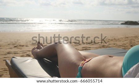 Young woman lying on sunbed by the sea and tanning. Female body on chaise-longue relaxing and enjoying during summer vacation on empty sandy ocean beach. Girl in bikini on a resort. Close up