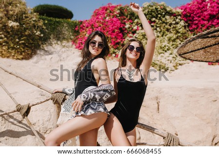Attractive smiling young woman posing with hand up standing near the best friend in trendy swimsuit. Gorgeous girls in stylish sunglasses having fun on summer resort dancing under the sun