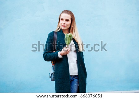 Stylish trendy woman wearing turquoise  green coat with the bouquet of flowers in her hands is standing in front of the blue wall.