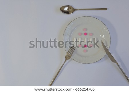 Pink, purple, white pills on a white plate  with fork, knife and spoon
