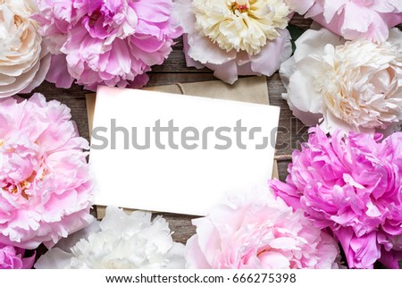 blank greeting card or wedding invitation and envelope in frame of tender peonies flowers over rustic wooden background. mock up. flat lay. top view with copy space