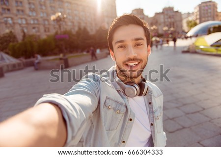 Selfie mania! Excited young guy is making selfie on a camera. He is wearing casual trendy wear and big modern headphones, on a walk in spring town outdoors Royalty-Free Stock Photo #666304333