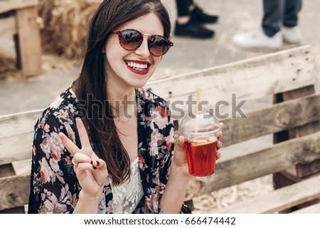 stylish hipster woman in sunglasses with red lips drinking lemonade. boho girl holding cocktail and smiling, showing peace sign. summertime. summer vacation travel. space for text