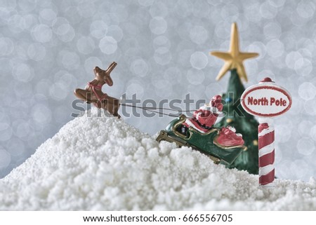 Small metal Christmas figurines of Santa, his sleigh, and a reindeer on fake snow. A North Pole sign and a Christmas tree faded into the silver bokeh background to finish the scene. 