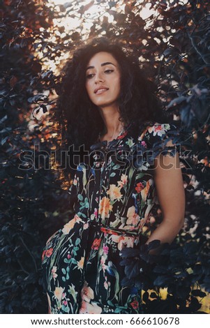 A model look woman with long dark curly hair wearing colorful summer dress is standing in the bushy area and looking at the camera. Photo of a caucasian brunette female standing beside the flowers.