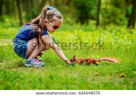 A little girl in denim overalls, a T-shirt, sneakers and glasses feeds a small red squirrel from her hand.