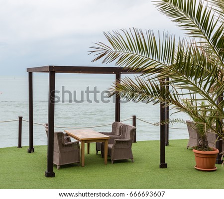 Wooden tables and wicker chairs under a canopy overlooking sea. Palm tree leaves