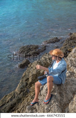 Young asian man traveler and photographer sitting on the rock cliff near tropical sea. Summer holiday or vacation travel. Relaxation and carefree concepts