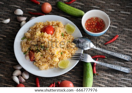 fried rice in the white plate with chili sauce in while cup,tomato,garlic,red chili,cucumber,fork and spoon on wooden.