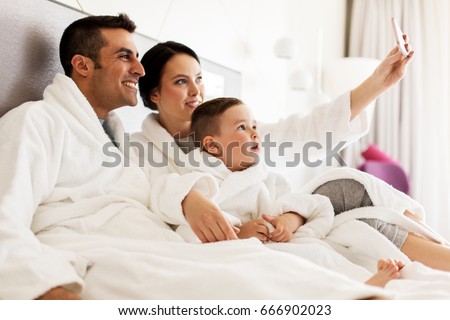 people, family and technology concept - happy mother, father and little boy in bathrobe taking selfie by smartphone in bed or hotel room