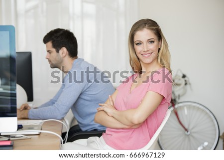 Shot of a young casual woman with arms crossed sitting at desk in the office while looking at camera and smiling. 